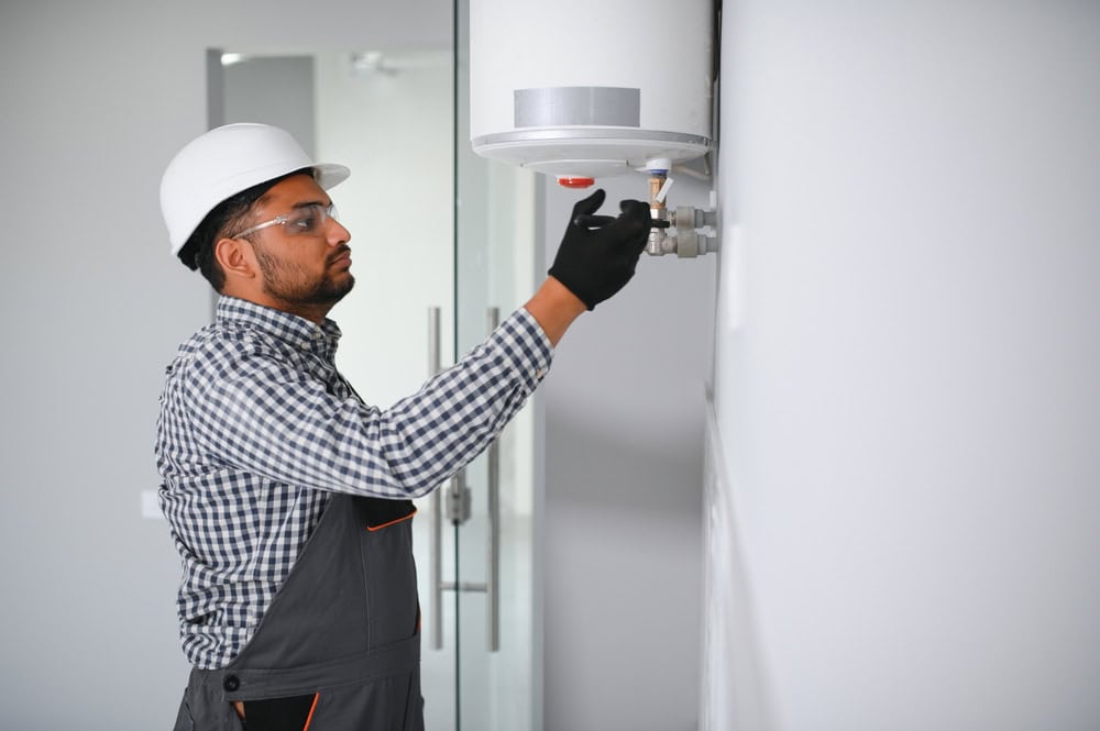 A man in a hard hat and safety glasses is adjusting a control on a wall-mounted cylindrical device, possibly a water heater. He is wearing a checkered shirt and overalls, and the room has white walls with a visible door.